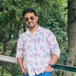 Raj nath joshi wearing sunglasses and a floral shirt stands outdoors in front of green trees, leaning casually against a railing and smiling at the camera.